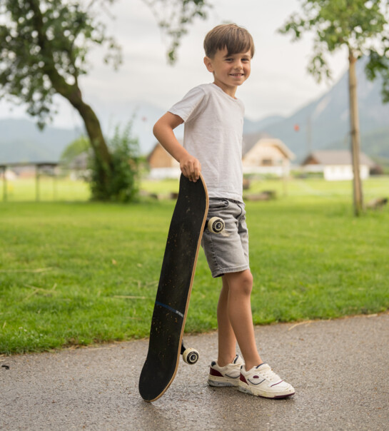 Bub steht, mit Skateboard in der Hand, auf der Straße und lächelt in die Kamera.